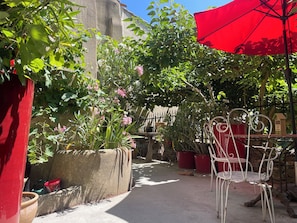 Un étroit sentier extérieur bordé de plantes en pot et surmonté d’un parasol rouge.