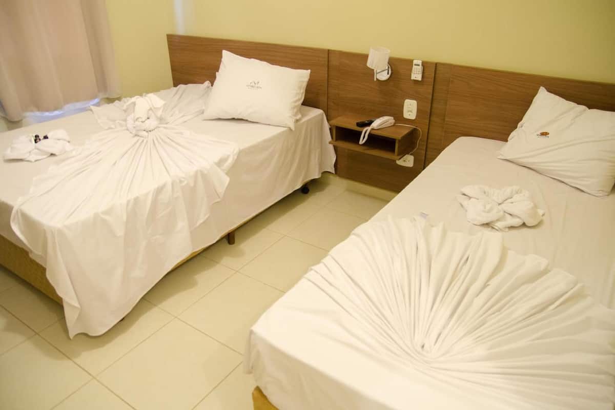 Two neatly made beds with white linens and towels arranged in a fan shape, a wooden headboard, a bedside telephone, and a small shelf with a phone and a book.