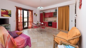 A living room with a sofa, a dining table with chairs, a kitchen area with red cabinets, and a balcony with a sliding door. - I tre Golfi Nespolo (San Vito Lo Capo)