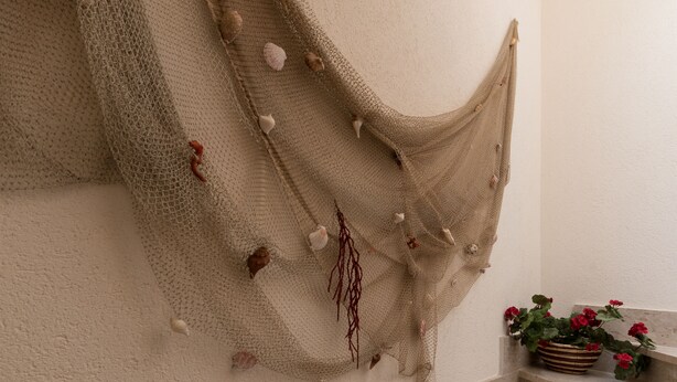 A beige net hanging on a textured wall with seashells and dried seaweed decorations. - I tre Golfi Nespolo (San Vito Lo Capo)