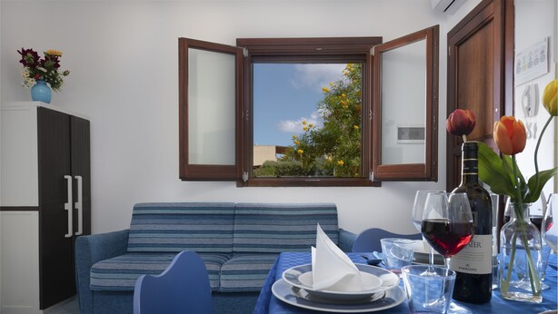A dining area with a table set for two, a sofa, and a window with an open shutter. - I tre Golfi Paprica (San Vito Lo Capo)