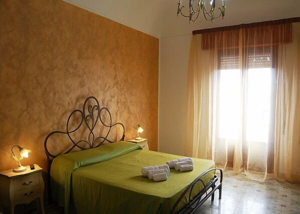 A bedroom with a green bedspread, two bedside lamps, a chandelier, and a window with sheer curtains.