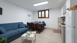 A modern living room with a blue sofa, a dining table set for six, and a kitchen area with a refrigerator and cabinets.