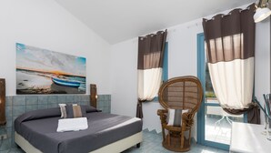 Une chambre à coucher avec un lit, une chaise en osier, un tableau représentant un bateau accroché au mur et une vue par la fenêtre.