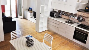 Une cuisine moderne avec des meubles blancs, un coin repas doté d’une table et de chaises blanches, et un canapé noir dans l’espace salon.