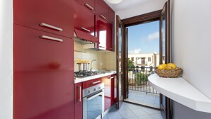 A compact kitchen with red cabinets, a stove, and a sink. A balcony with a wicker basket of lemons is visible. - I tre Golfi Zenzero (San Vito Lo Capo)