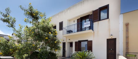 A two-story building with a balcony, a door with a sign, and a small garden with yellow flowers.
