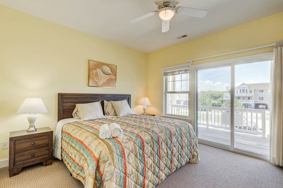 A bedroom with a bed, two bedside tables with lamps, a ceiling fan, a large window with a view of buildings, and a painting on the wall.
