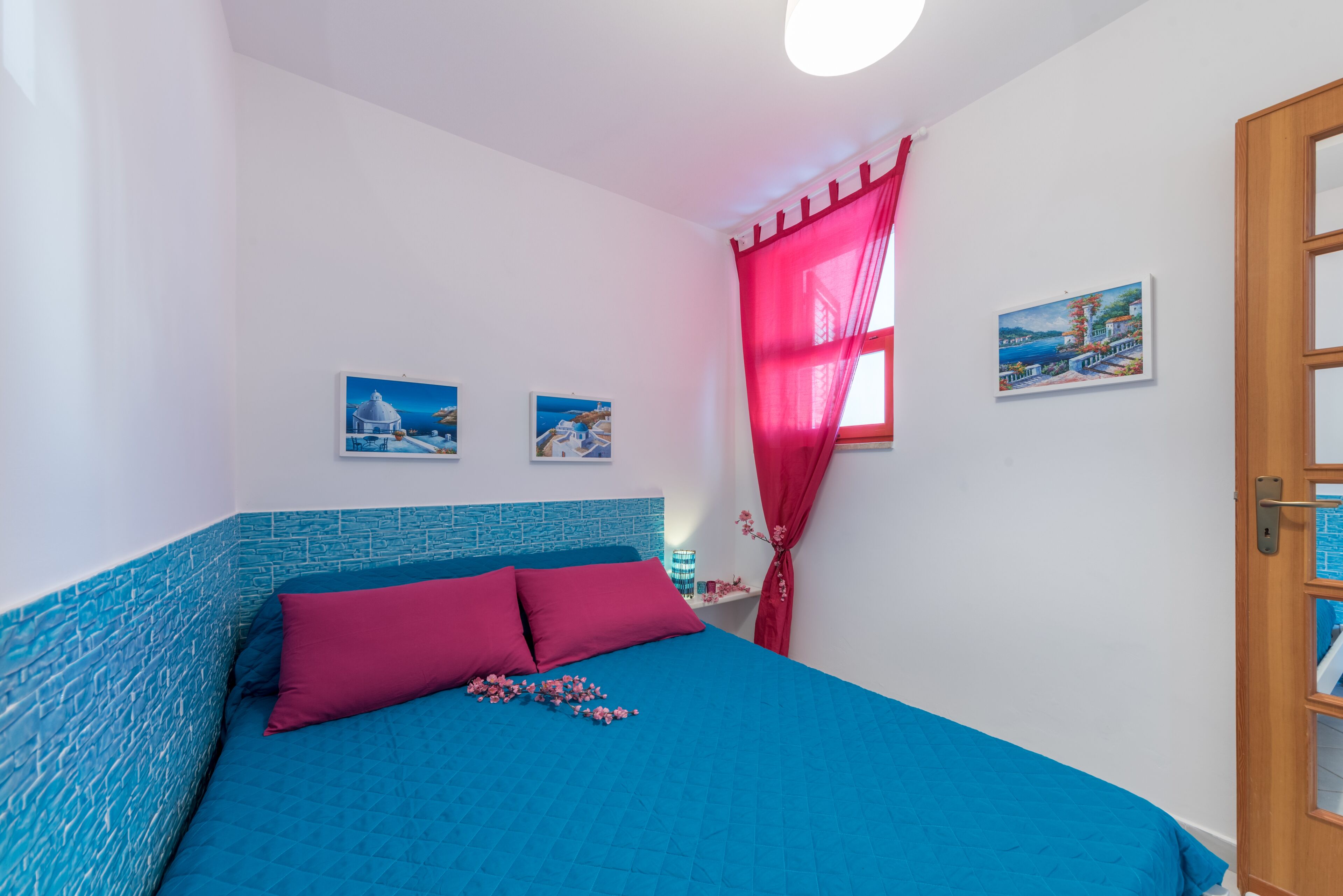 Une chambre avec une couvre-lit bleue, deux coussins roses, un rideau rouge et deux tableaux encadrés accrochés au mur.