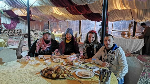 A group of people dining at a table with various dishes and utensils, under a tented roof with draped fabric.
