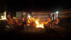 A group of people sitting around a campfire at night, with tents and a striped wall in the background.