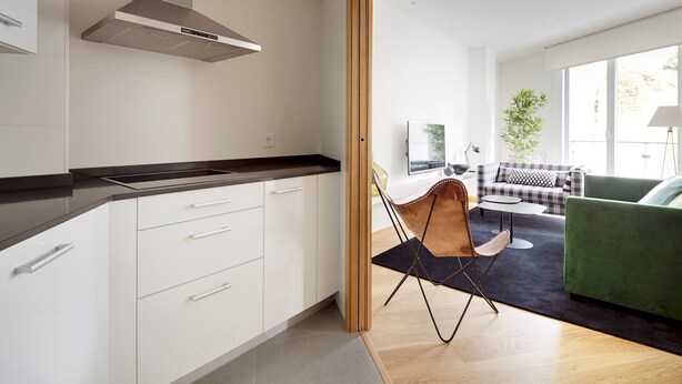 A modern kitchen with white cabinets and a black countertop, a dining area with a green sofa and a black and white checkered table, and a large window providing natural light. - Muinoa 1 by FeelFreeRentals (Donostia)