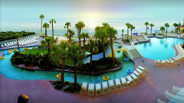 A pool area with lounge chairs and palm trees, overlooking a beach.