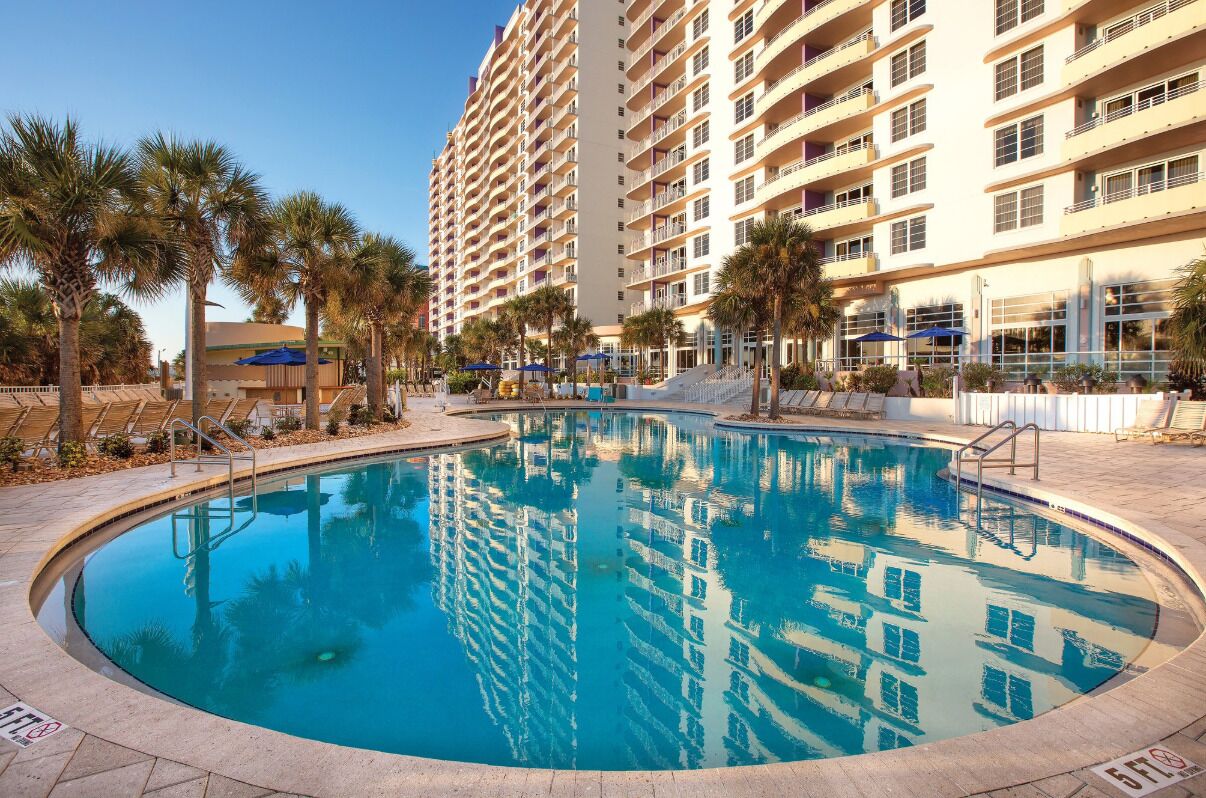 A swimming pool with a clear blue water, surrounded by palm trees and a tall building in the background.