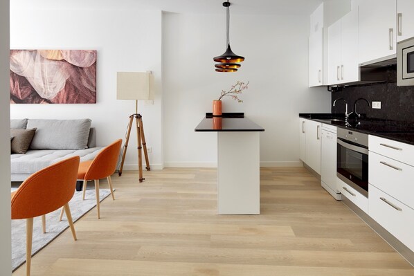 A modern kitchen with white cabinets, a black countertop, and a central island. There is a dining area with a gray sofa and orange chairs.