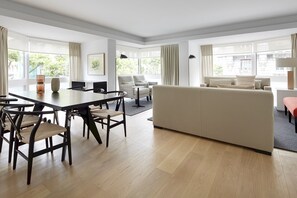 A modern living room with a large sofa, a dining table with chairs, and a view of the outdoors through large windows.