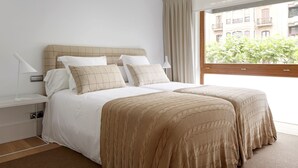 A double bed with a beige headboard and white bedding, a bedside table with a lamp, and a view of a building through a window with curtains. - Enbata by FeelFreeRentals (San Sebastian)