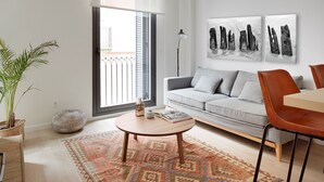 Uma sala de estar moderna com um sofá cinza, uma mesa de centro redonda de madeira, um tapete estampado, uma planta em vaso e uma grande janela com vista.