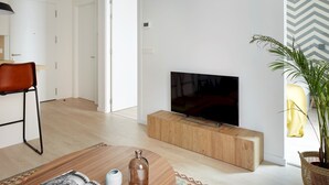 Uma sala de estar moderna com uma televisão de tela plana sobre um suporte de madeira, uma área de jantar com uma poltrona de couro marrom, e um vaso de planta perto da janela.