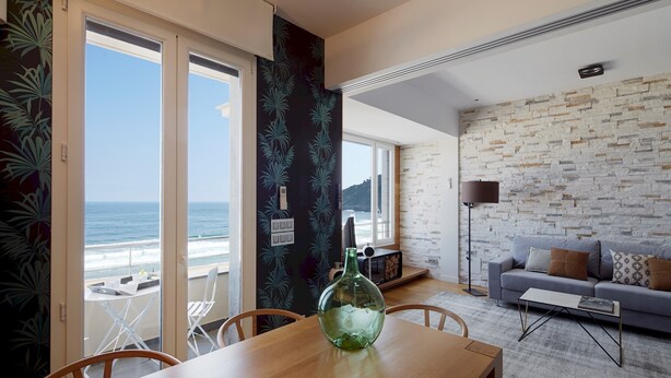 A room with a wooden table, a green vase, a grey sofa, and a view of the ocean. - Kantauri by FeelFreeRentals (Donostia)