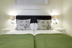 Uma cama de casal bem arrumada com roupa de cama verde, duas almofadas iguais e duas luminárias de cabeceira sobre mesas de cabeceira brancas.