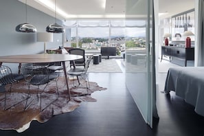 Quarto de hotel moderno com área de refeições, uma janela grande com vista para a cidade, uma cama e um banheiro.