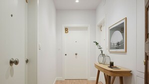 Un couloir doté d’un plancher en bois, de murs blancs et de portes marquées des lettres A et B. On y trouve une table en bois sur laquelle sont posés un vase et un tableau encadré accroché au mur.
