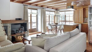 Un salon chaleureux avec une cheminée, un téléviseur et un canapé. Il y a également un coin repas avec une table et des chaises. La pièce est dotée de poutres en bois au plafond et de grandes fenêtres.
