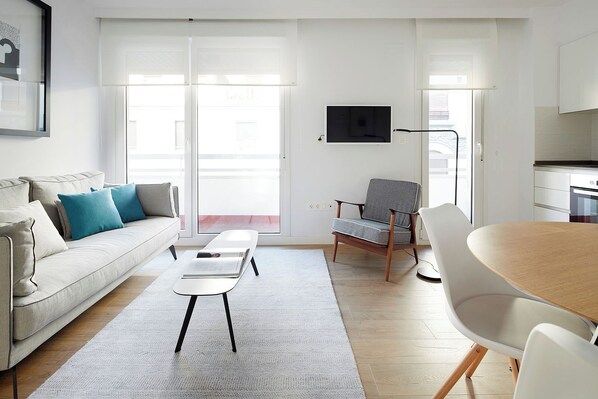 A modern living room with a sofa, a coffee table, a dining table, and a chair. There is a TV mounted on the wall and a window with a view of the outside. - San Francisco 2 by FeelFree Rentals (Donostia)