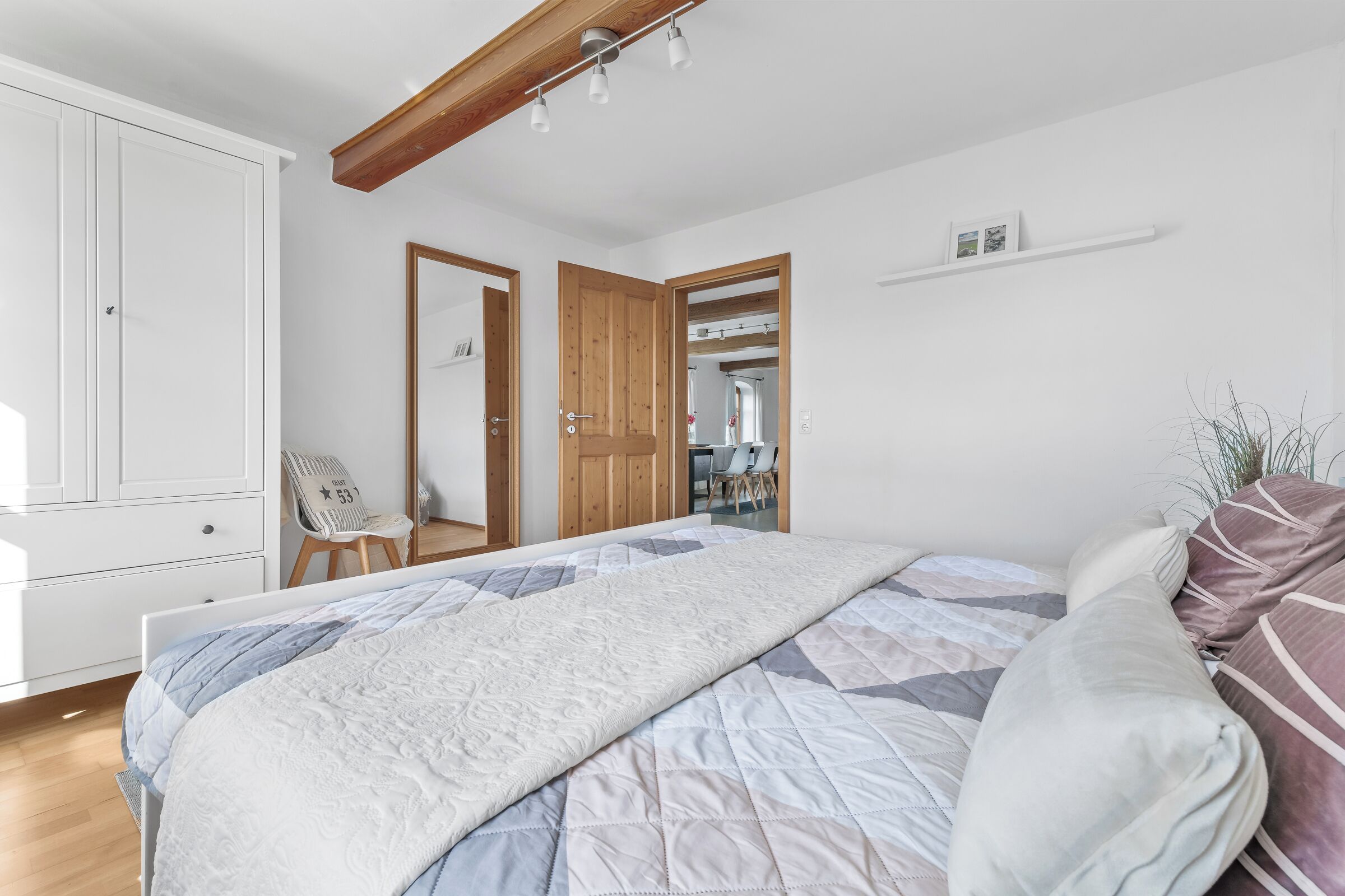 A bedroom with a large bed, wooden ceiling beams, a white wardrobe, and a small desk with a chair.
