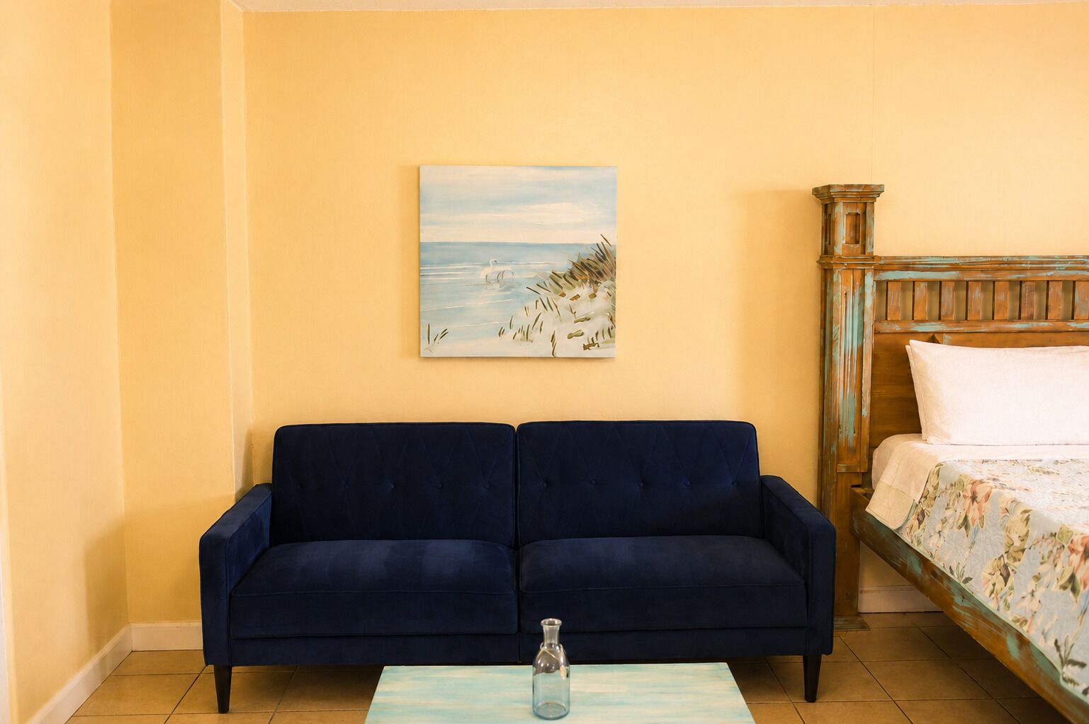 Quarto com um sofá azul, uma cama com roupa de cama floral, um quadro de uma praia e uma pequena mesa com um vaso de vidro.