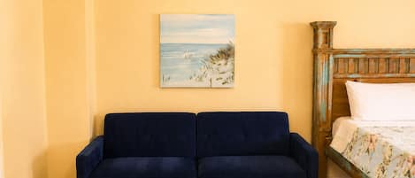 Quarto com um sofá azul, uma cama com roupa de cama floral, um quadro de uma praia e uma pequena mesa com um vaso de vidro.
