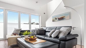 Een moderne woonkamer met een zwarte leren bank, een houten salontafel met een fruitschaal, en een groot raam met uitzicht op zee.