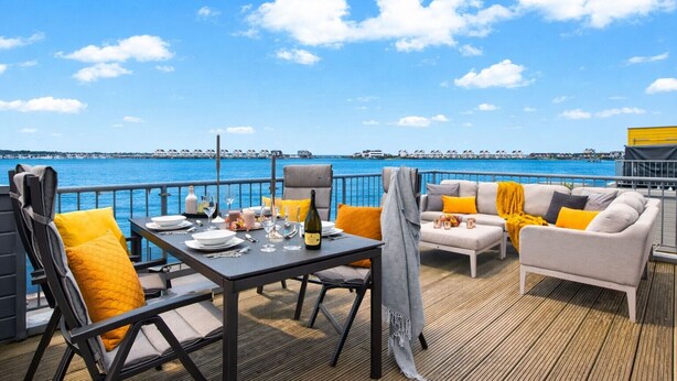 Een terras met een eettafel, een bank en stoelen, met uitzicht op een waterpartij en gebouwen in de verte.