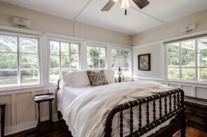 A bedroom with a large bed, multiple windows, a ceiling fan, and a nightstand.