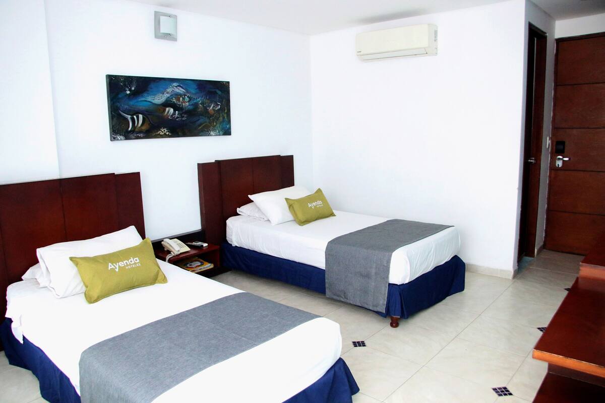 Una habitación de hotel con dos camas, un cuadro en la pared, aire acondicionado y una puerta.