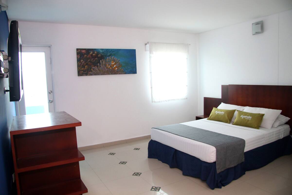 Una habitación de hotel con cama, escritorio, un cuadro en la pared y ventana con cortinas.