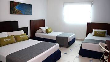 Ein Hotelzimmer mit drei Betten, jedes mit einem Kopfkissen mit dem Aufdruck "Ayenda Hotels". An der Wand hängt ein Gemälde, es gibt ein Fenster mit Jalousien und einen Sessel in der Ecke.