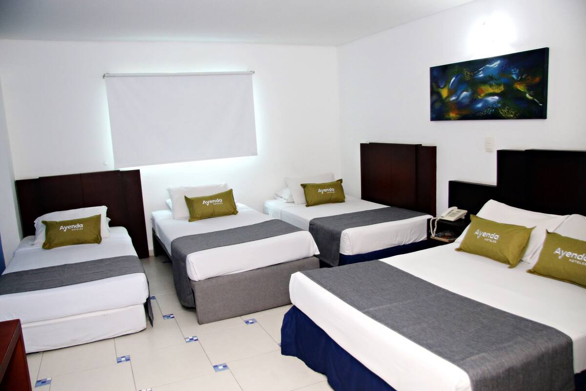 Habitación de hotel con tres camas, cada una con un almohadón con la etiqueta "Ayenda". Hay un cuadro en la pared y una ventana con persiana.