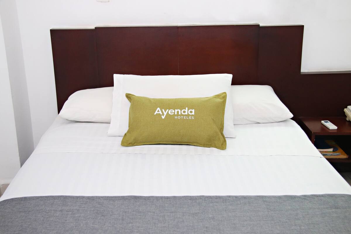 Una cama bien hecha con ropa blanca y una almohada verde con el texto 'Ayenda HOTEL'.