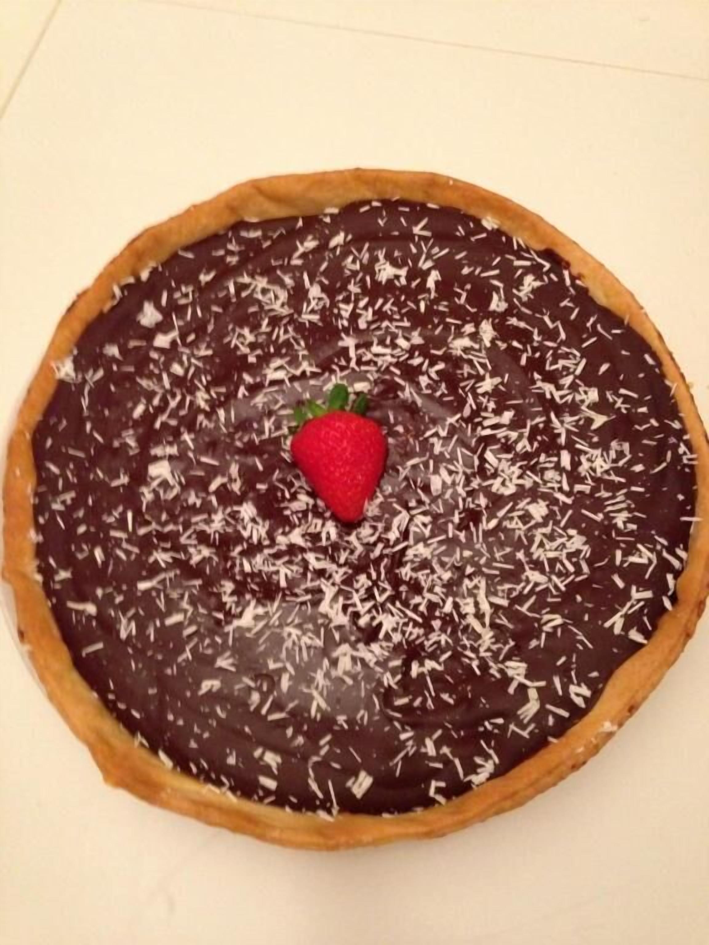 A round chocolate tart with a strawberry on top, placed on a white surface.