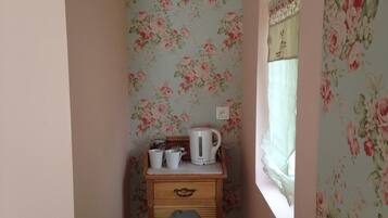 Una esquina de habitación con papel tapiz floral, una pequeña mesa de madera con un hervidor, tazas y un tarro, y una ventana con cortina.