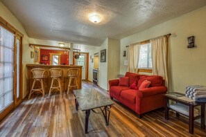 A cozy living room with a red sofa, wooden floor, and a dining area with wicker chairs. - L'Auberge Des Oiseaux Chantants (Willeman)