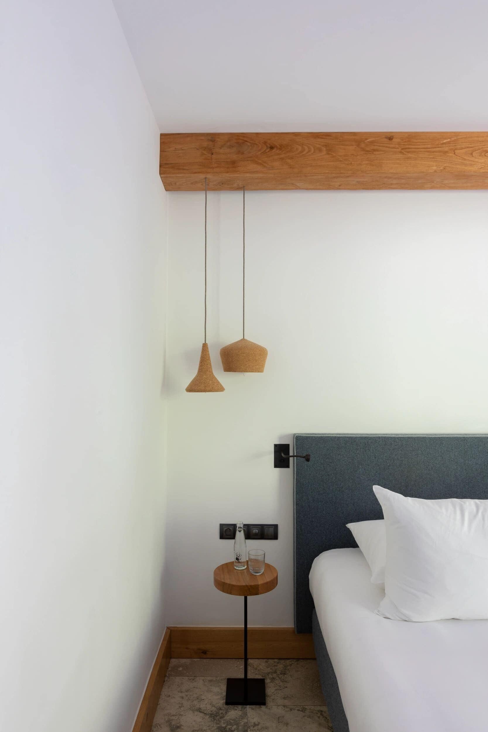 A bedroom with a wooden headboard, a bed with white bedding, a small bedside table with a glass and a candle, and two pendant lights.