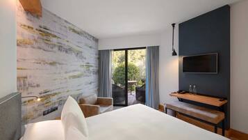 A modern hotel room with a large bed, a flat-screen TV mounted on the wall, a small table with a bench, and a balcony with outdoor seating.