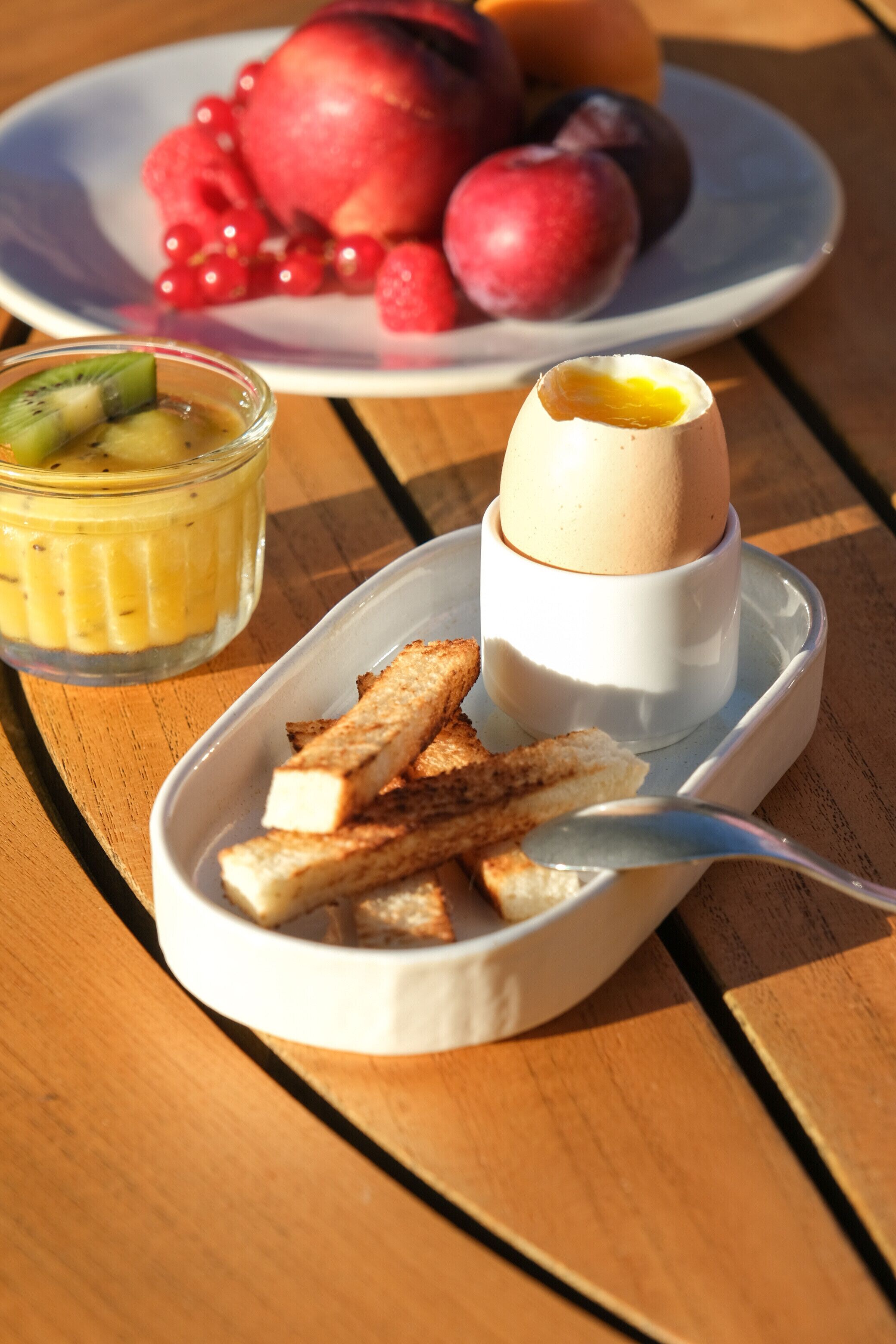A dessert plate with a poached egg, a glass of fruit salad, and toasted breadsticks on a wooden table.