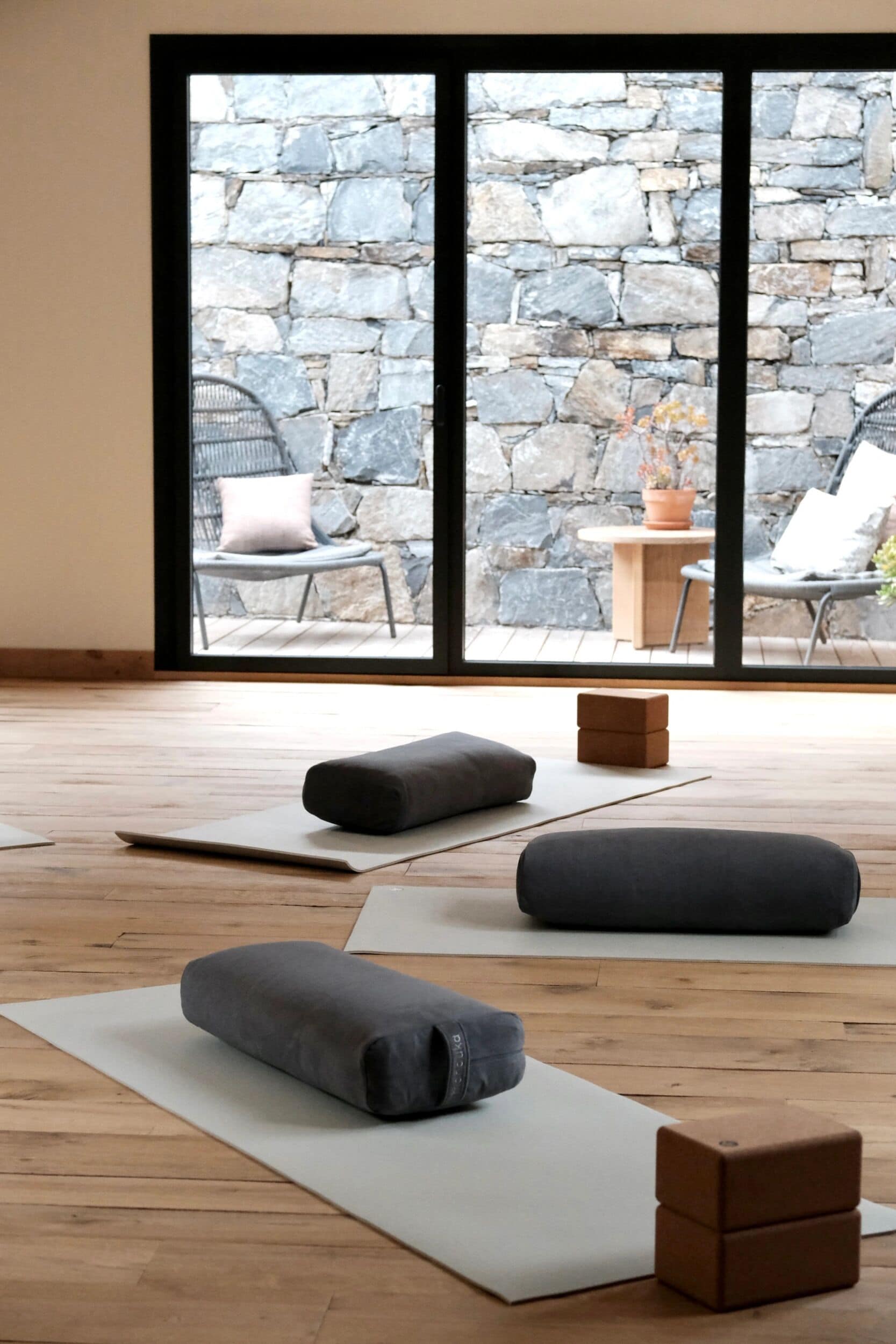 Ein Yoga-Raum mit Holzboden, Yogamatten und einer Steinsäule, die durch ein großes Fenster sichtbar ist.