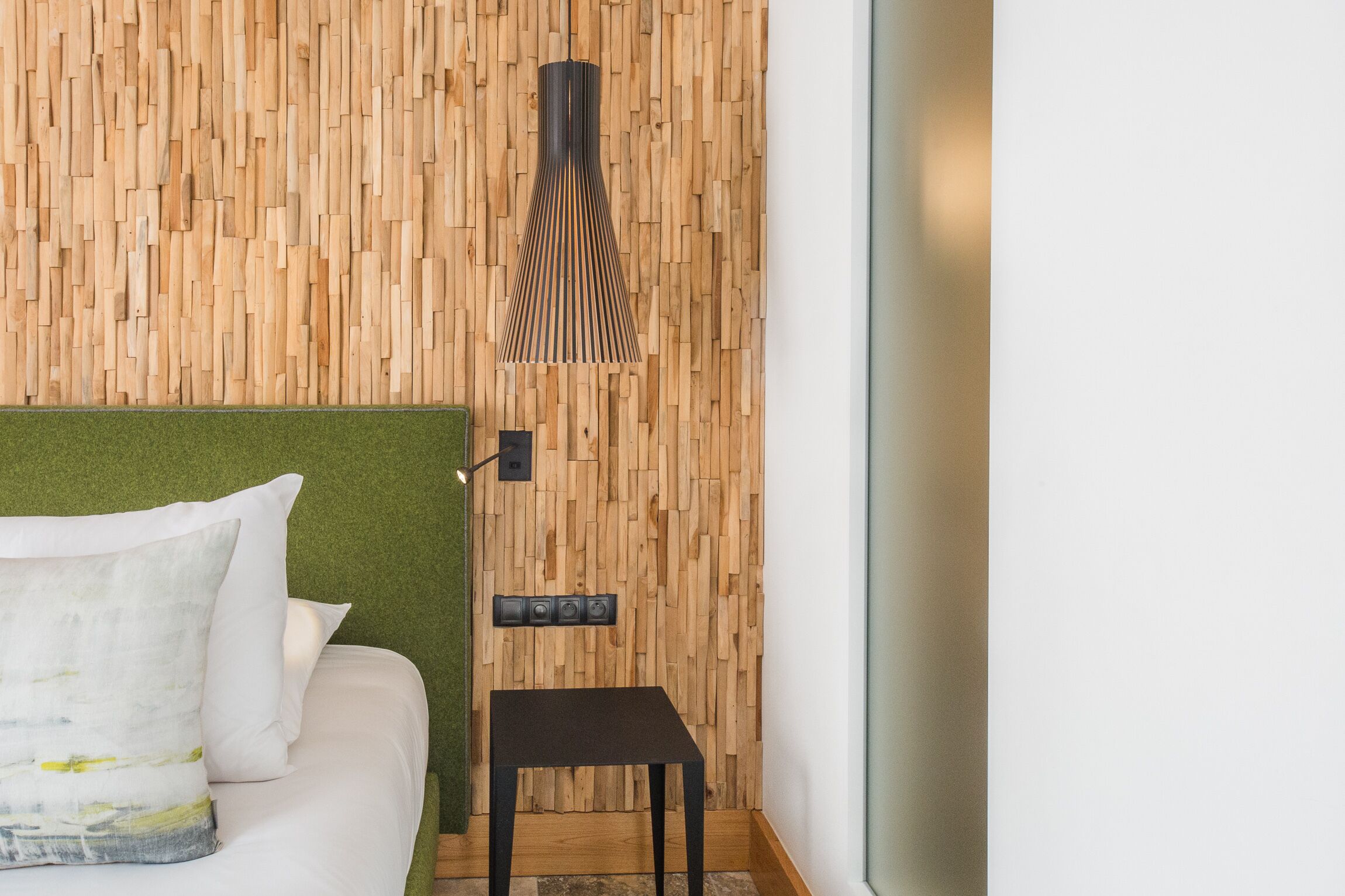 A room with a wooden wall panel, a bed with white bedding, a small black table, and a wall-mounted light fixture.