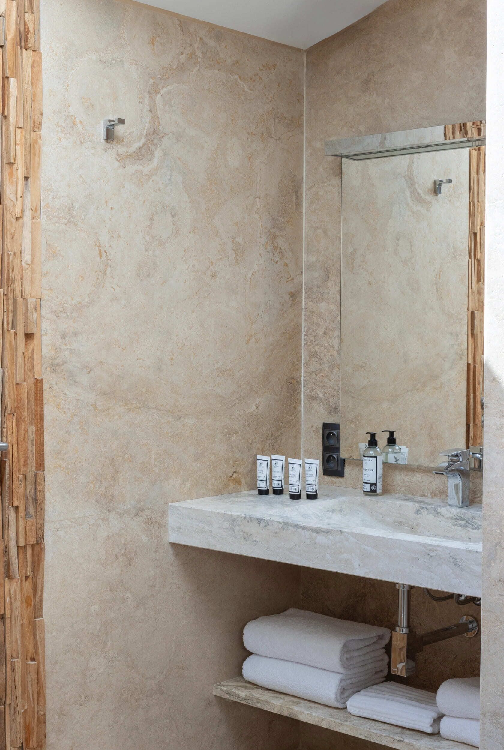 A bathroom with a marble wall, a wooden panel on the left, a white countertop with toiletries, and a towel rack below.