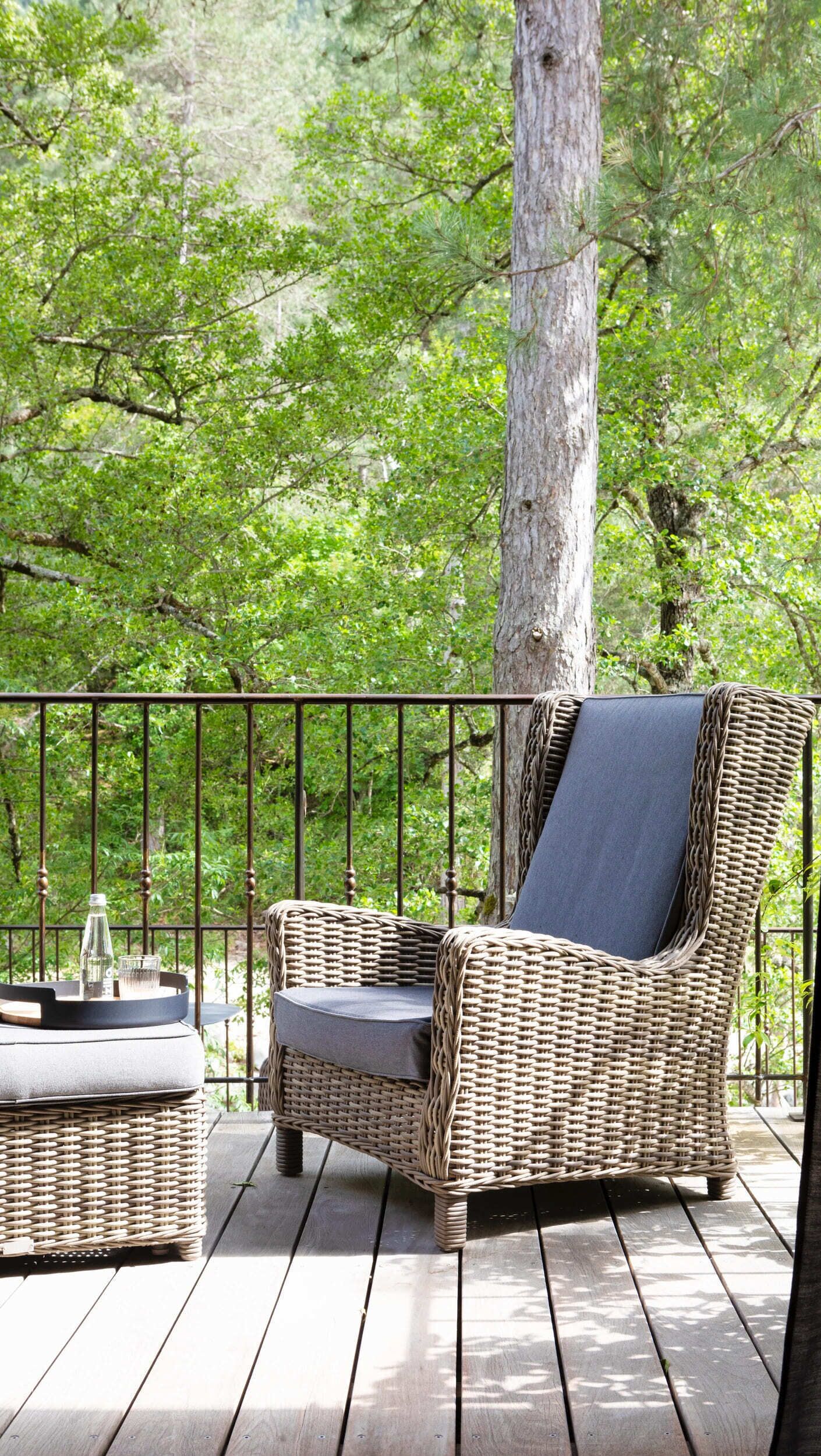 A wicker chair with a cushion on a wooden deck, surrounded by a metal railing, with a tree in the background.